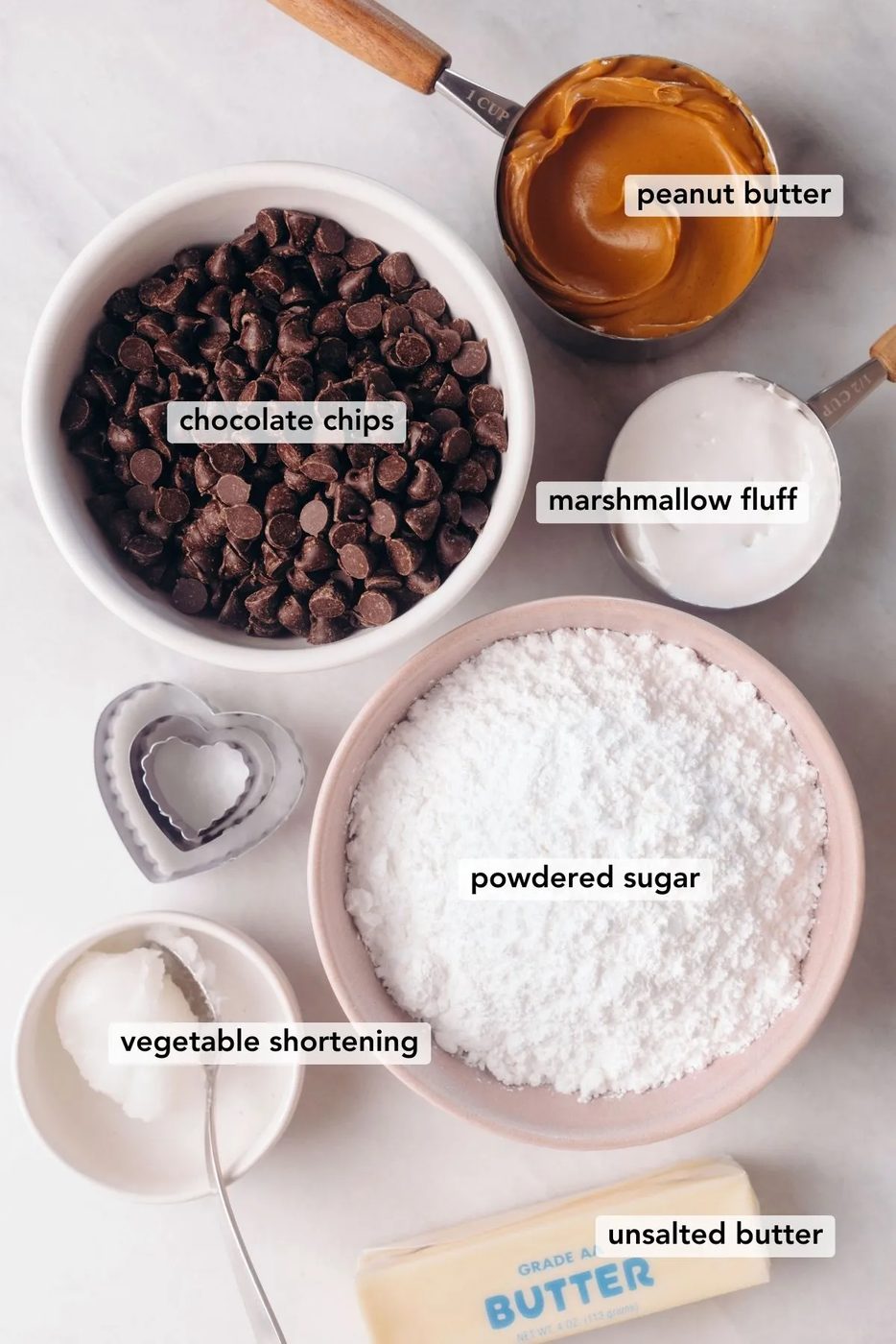 Ingredients needed for chocolate covered peanut butter eggs with three heart-shaped cookie cutters on a white marbled background.