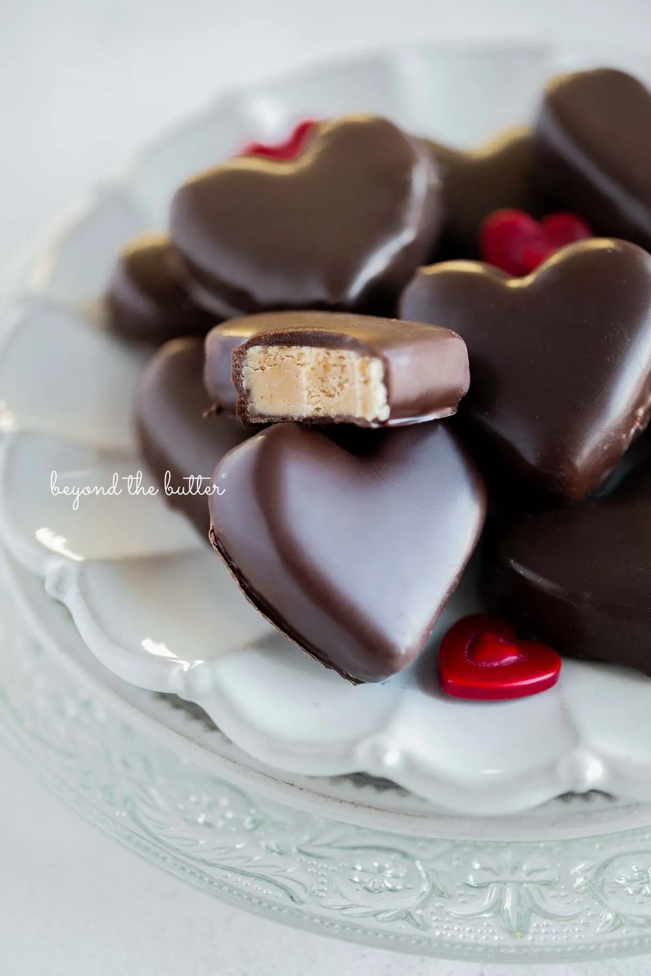 Chocolate covered peanut butter hearts on a white ceramic dessert plate on white background with bite taken out of center peanut butter heart.