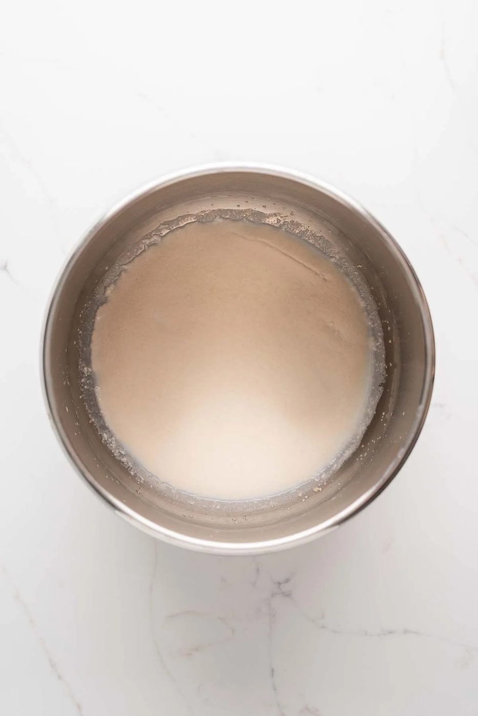 Proofing yeast in warm milk and sugar in a large stainless steel Ankarsrum mixing bowl on a white marbled background.
