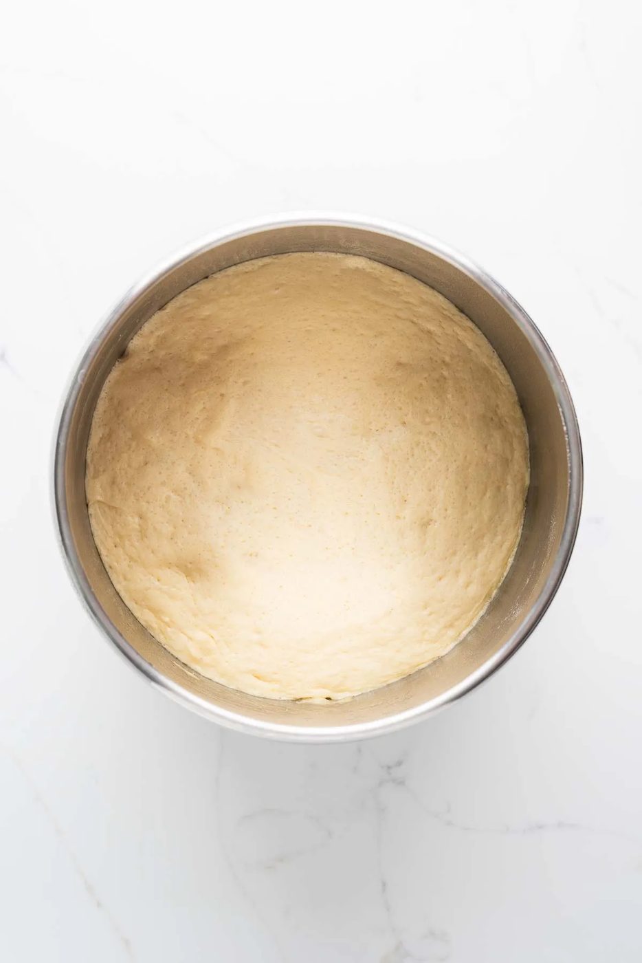 First rise of croissant dough in a stainless steel mixing bowl on a white marbled background.