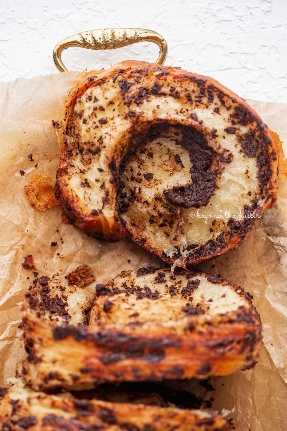 Sliced chocolate croissant loaf placed on brown parchment paper lined gold tray and white background.