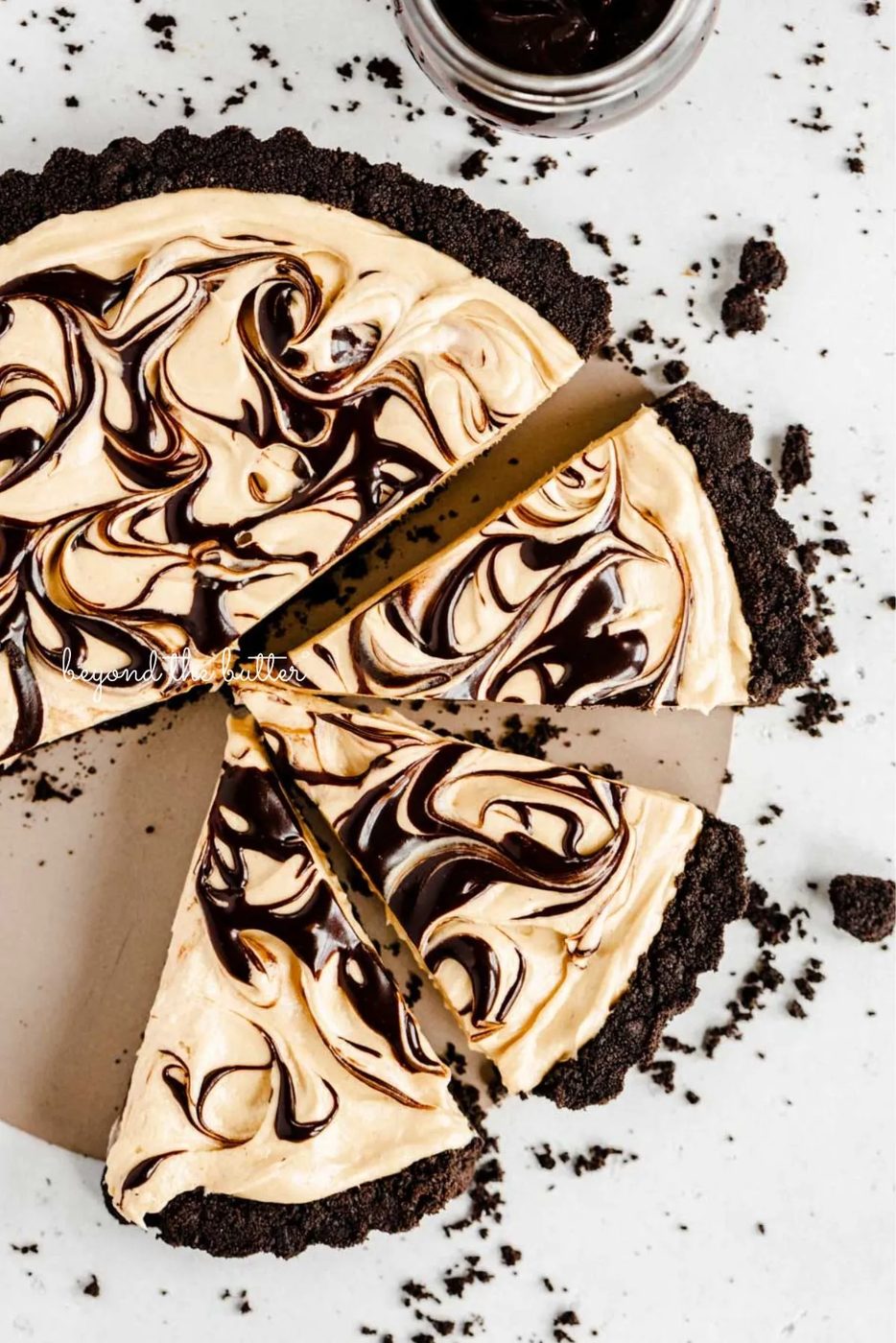 Round chocolate peanut butter tart swirled with chocolate syrup and cut into slices with a bowl of chocolate sandwich crumbs next to it on a white speckled background.