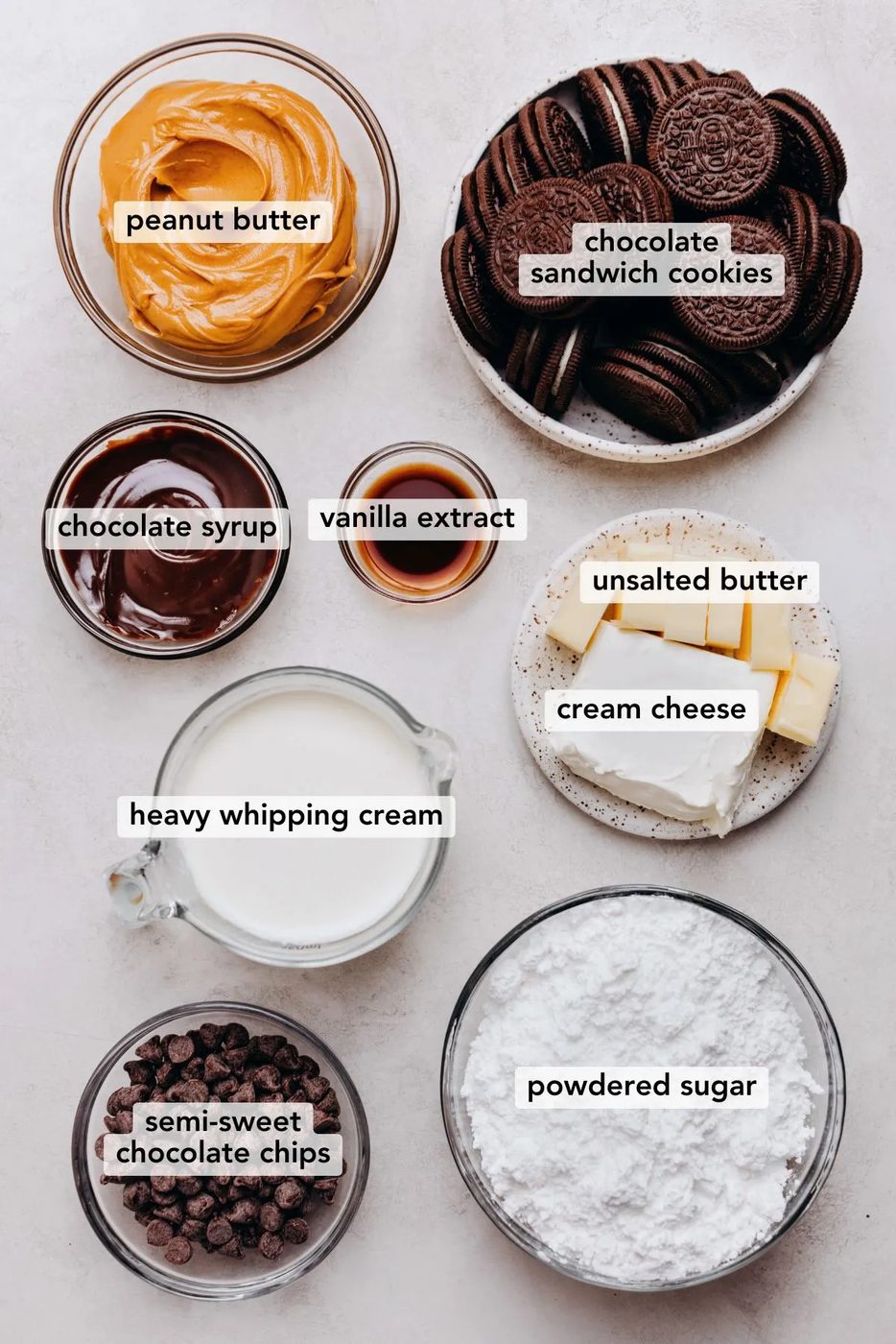 Ingredients in various size bowls labeled as peanut butter, chocolate sandwich cookies, chocolate syrup, vanilla extract, unsalted butter, cream cheese, heavy whipping cream, semi-sweet chocolate chips, and powdered sugar on a white marbled background.