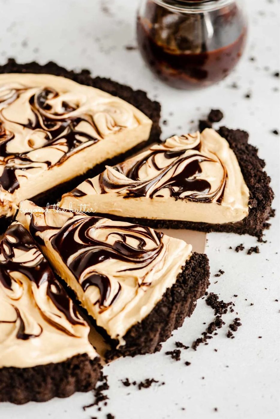 Round chocolate peanut butter tart swirled with chocolate syrup and cut into slices with a bowl of chocolate syrup next to it surround by a few chocolate sandwich cookie crumbs on a white speckled background.