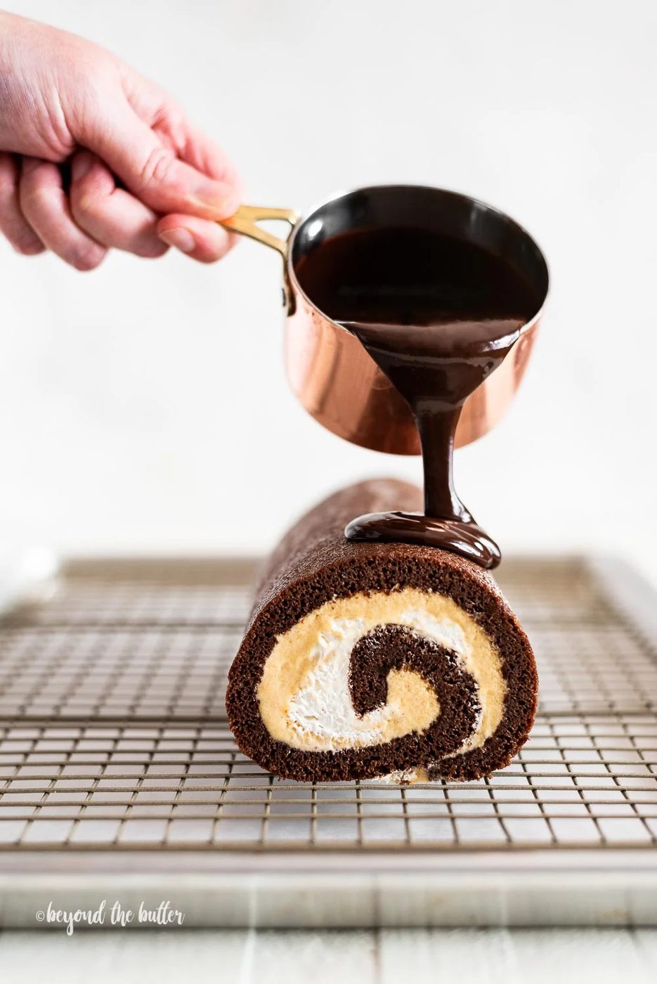 Pouring melted chocolate from small copper pot over chocolate pumpkin roll on wire cooling rack.