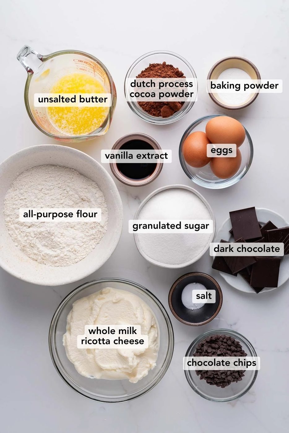 Bowls of different ingredients used to make chocolate ricotta cake on a white marbled background.