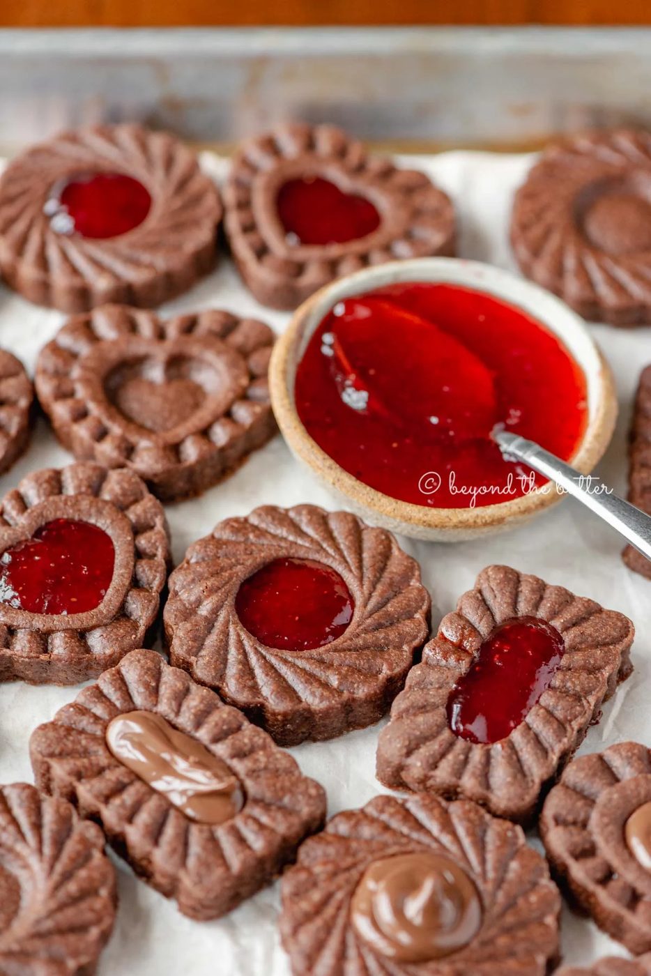 Parchment lined baking sheet full of chocolate thumbprint cookies with small bowl of strawberry jam and spoon | All Images © Sovuh