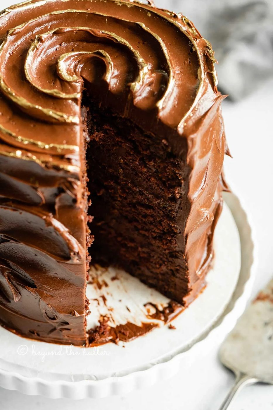 Angled closeup image of chocolate zucchini cake with gold decorative swirl on top and slice removed | All Images © Sovuh