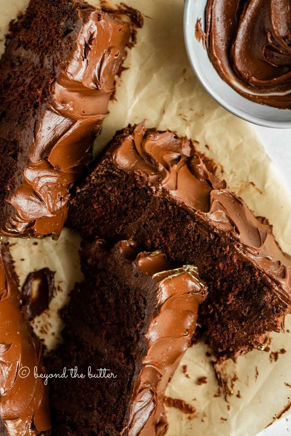 Overhead image of chocolate zucchini cake slices on parchment paper with a small bowl of frosting | All Images © Sovuh