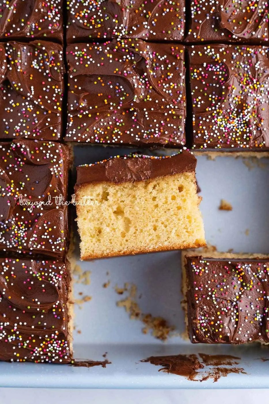 Slices of four egg yellow sheet cake with chocolate frosting and rainbow nonpareils in a blue baking pan with center slice placed on its side to see the tender cake and thick layer of frosting.