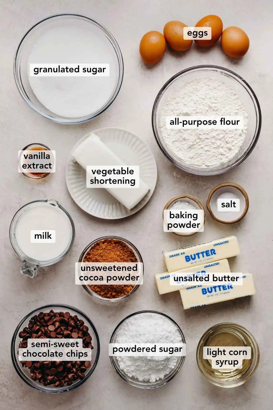 Ingredients in various size bowls labeled as granulated sugar, eggs, all purpose flour, vanilla extract, vegetable shortening, baking powder, salt, milk, unsweetened cocoa powder, unsalted butter, semi sweet chocolate chips, powdered sugar, and light corn syrup on a white marbled background.