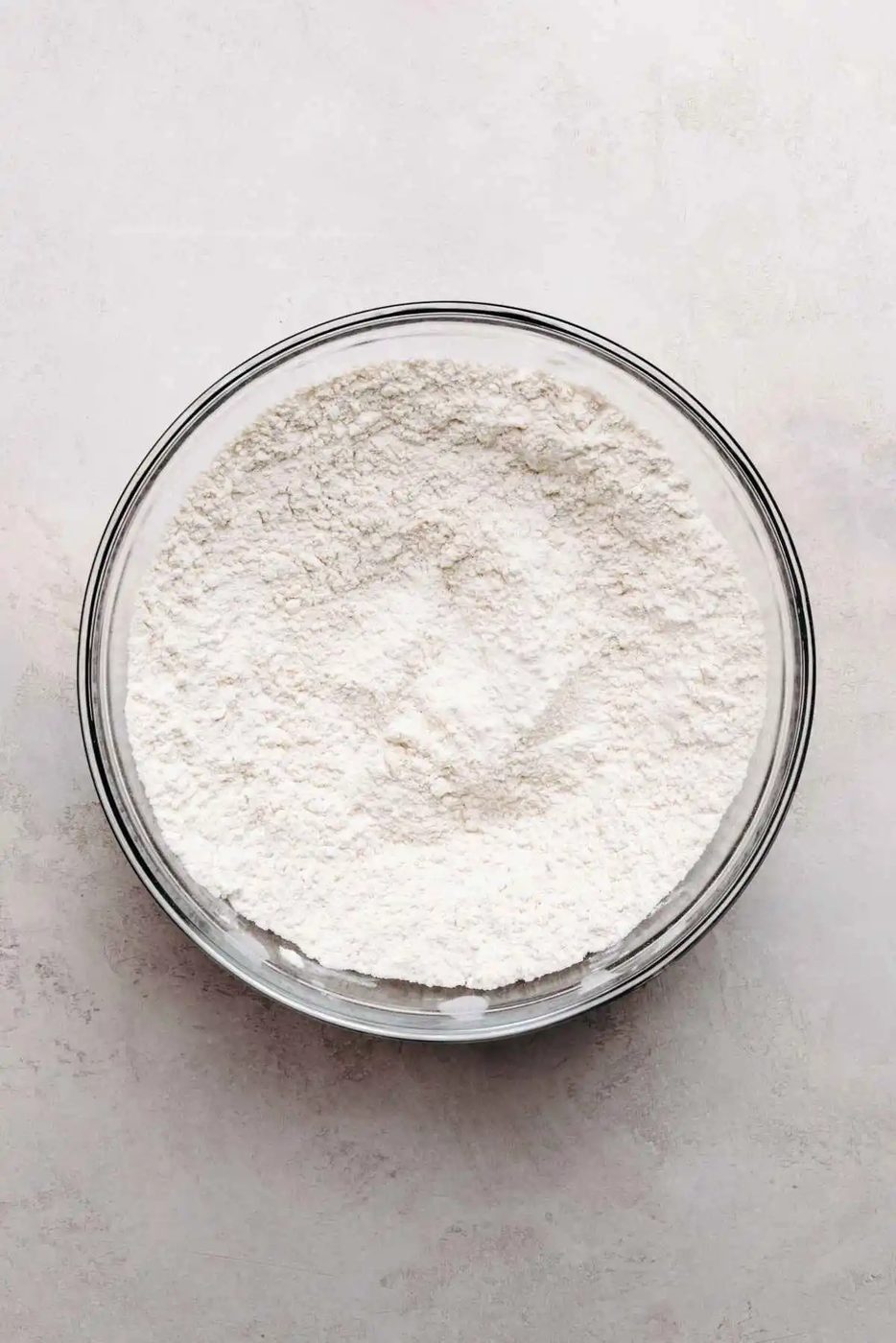 All purpose flour, baking powder and salt whisked together in a medium size glass bowl on a grey marbled background.