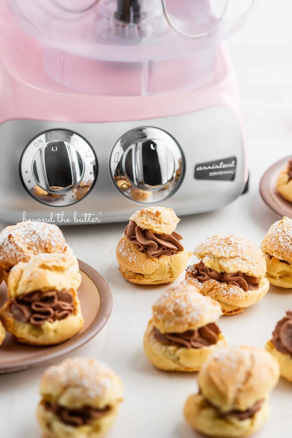 Cloudy pink Ankarsrum mixer with cream puffs filled with chocolate whipped cream and dusted with powdered sugar placed around it on pink dessert plates and white marbled background.
