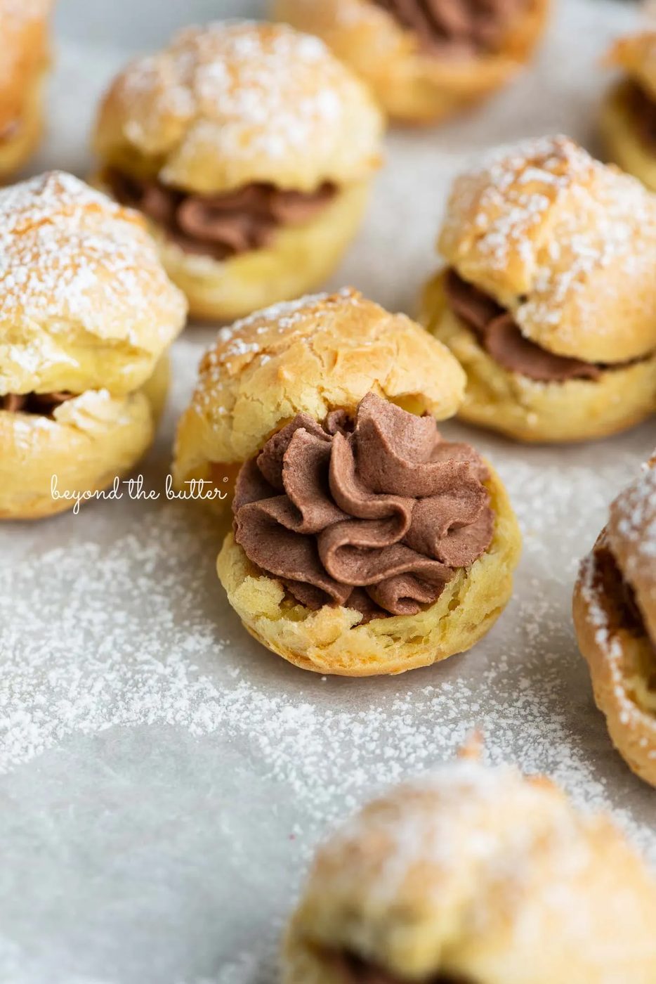 Cream puffs filled with chocolate whipped cream and dusted with powdered sugar on a parchment paper lined baking sheet.