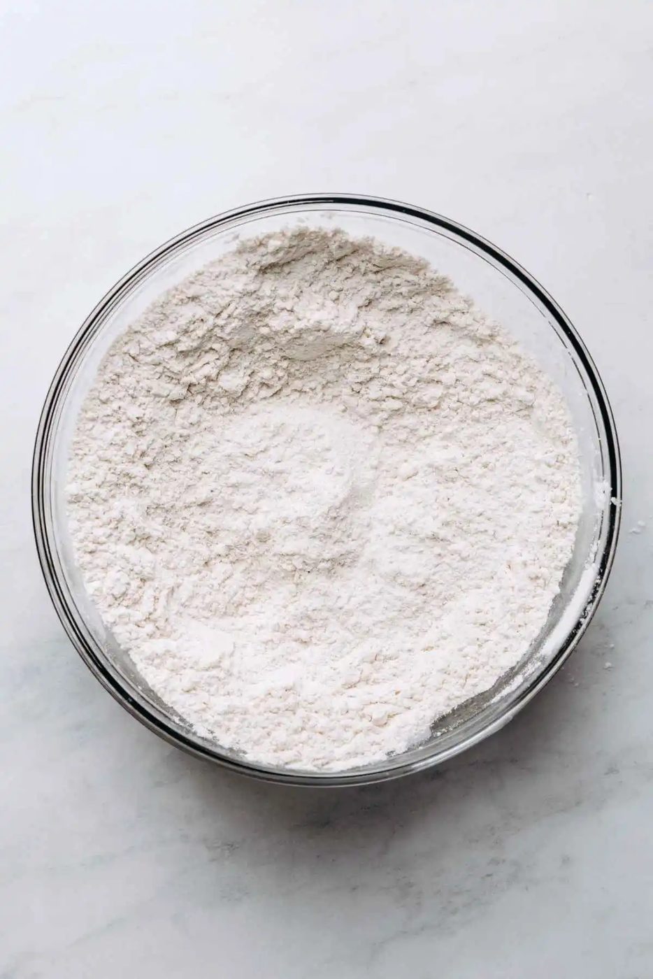 All purpose flour, baking soda, and salt whisked together in a glass mixing bowl on a white marbled background.