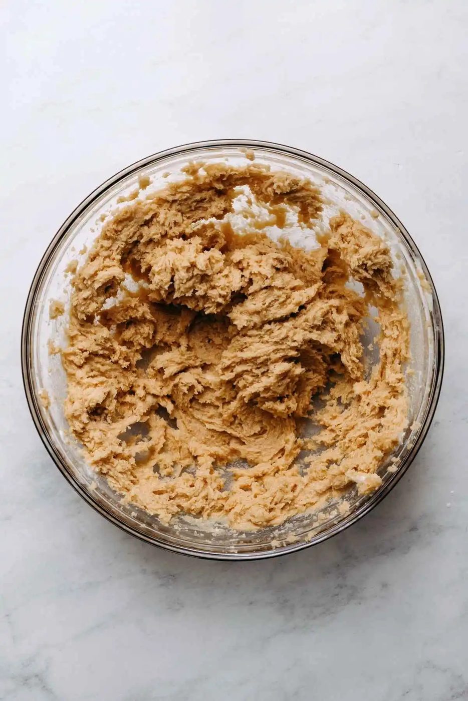 Unsalted butter and light brown sugar creamed together in a large glass mixing bowl on a white marbled background.