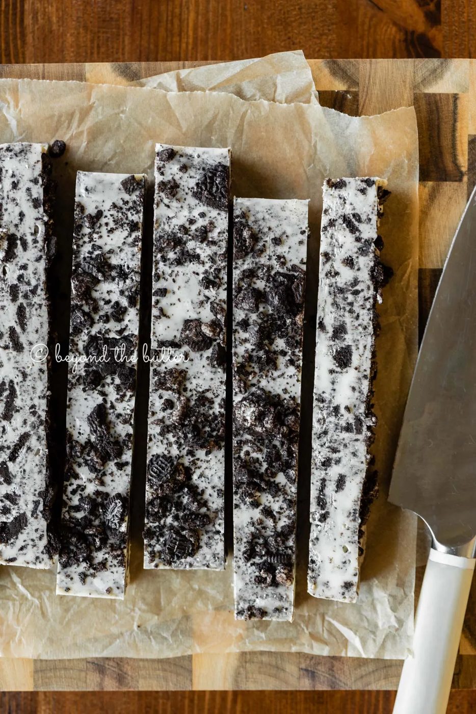 Bars of cookies and cream fudge on brown parchment paper and wood cutting board with knife on top | All Images © Sovuh