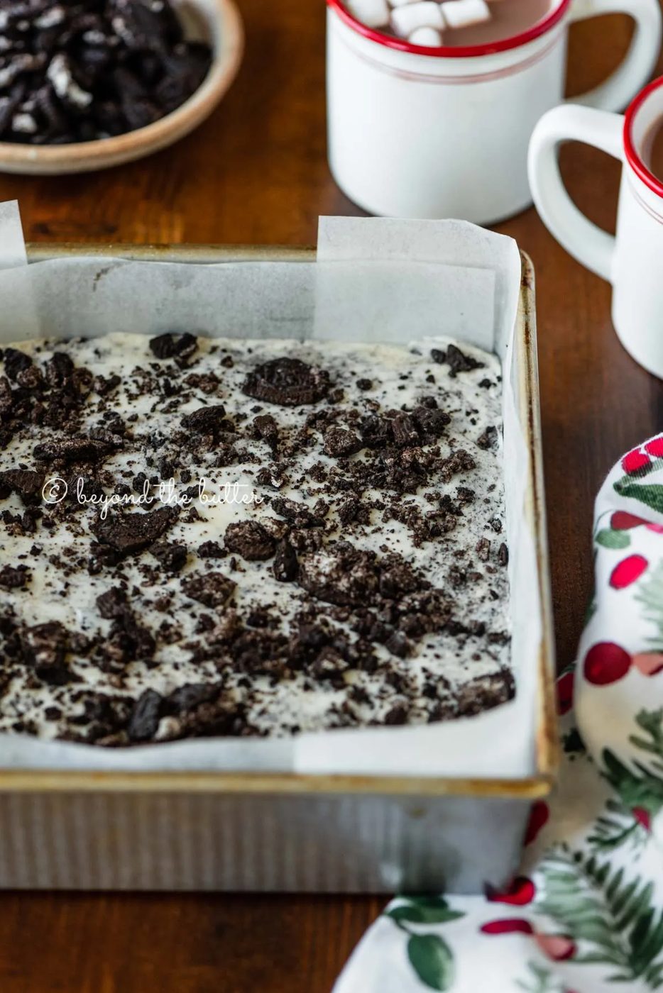 Angled image of cookies and cream fudge in prepared pan with holiday themed towel, mugs of hot chocolate, and crushed chocolate sandwich cookies nearby