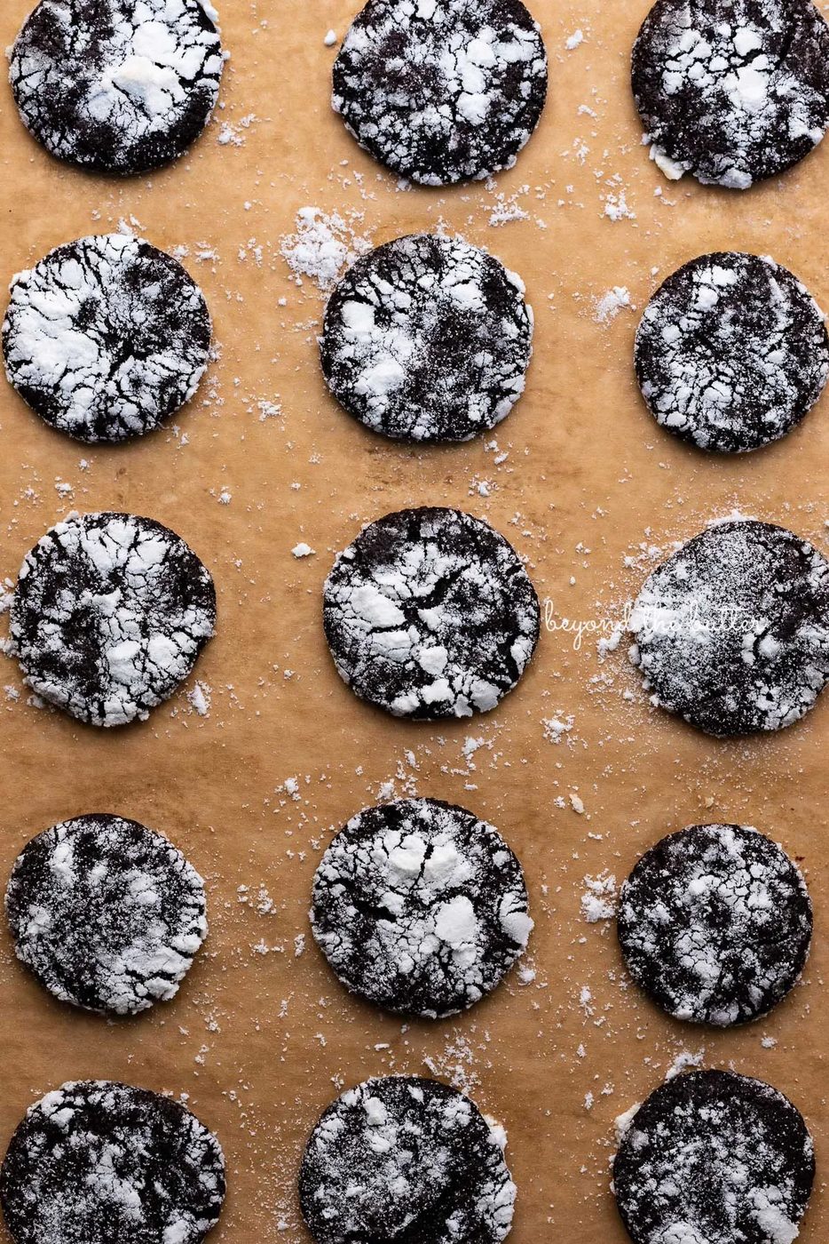 Dark chocolate crinkle cookies evenly spaced on a brown parchment paper lined baking sheet | © Sovuh