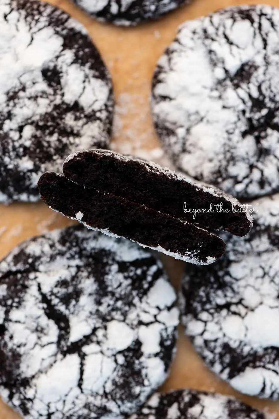 Split dark chocolate crinkle cookie to show the fudgy inside on parchment paper lined baking sheet | © Sovuh