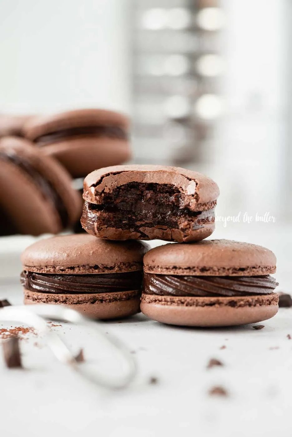 Small stack of chocolate macarons with a bite taken out of the top macaron with a bigger stack in the background with a sifter | All Images © Sovuh