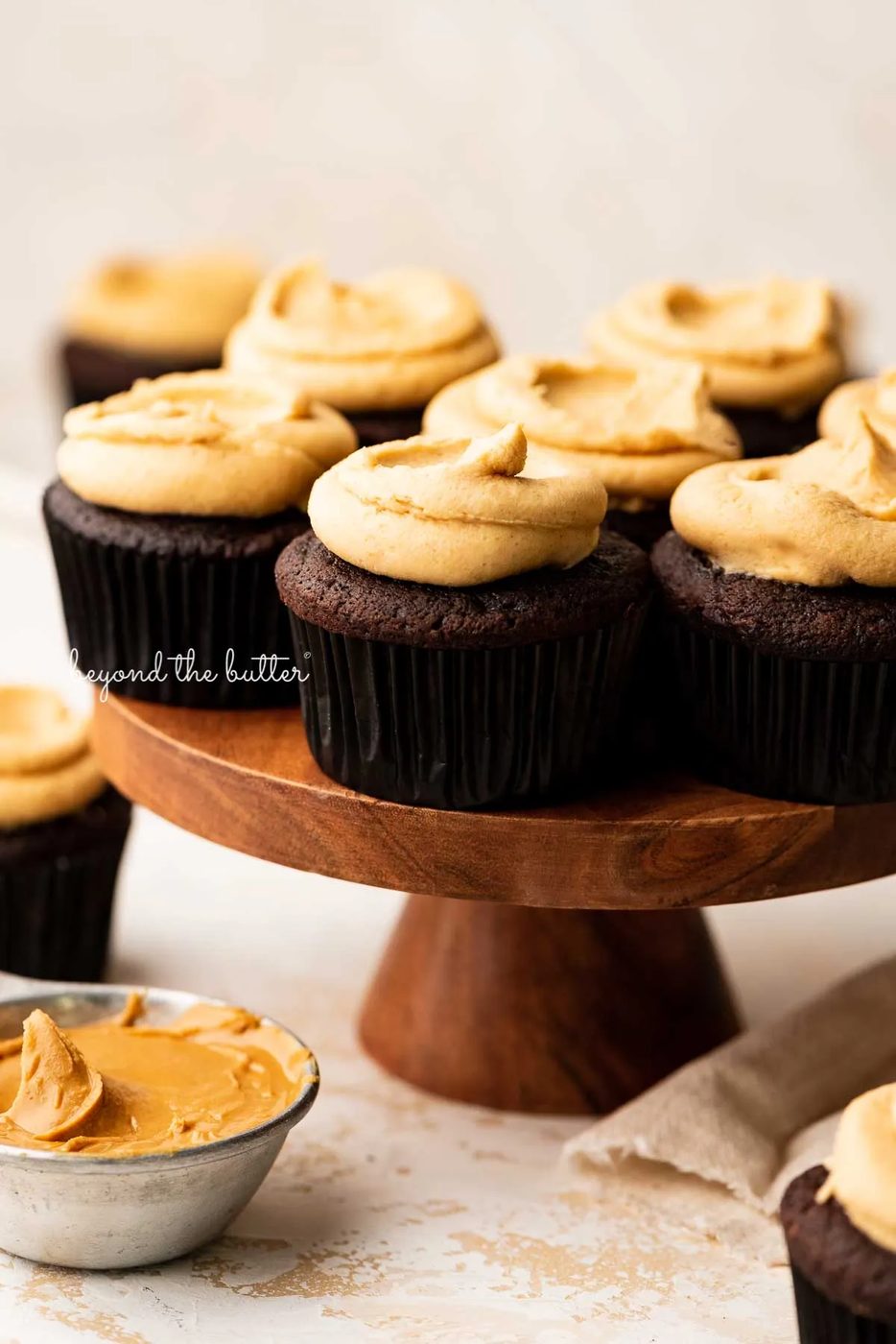 Wood cake stand with devil's food cupcakes surrounded by other cupcakes, peanut butter, and a napkin on a white background | © Sovuh