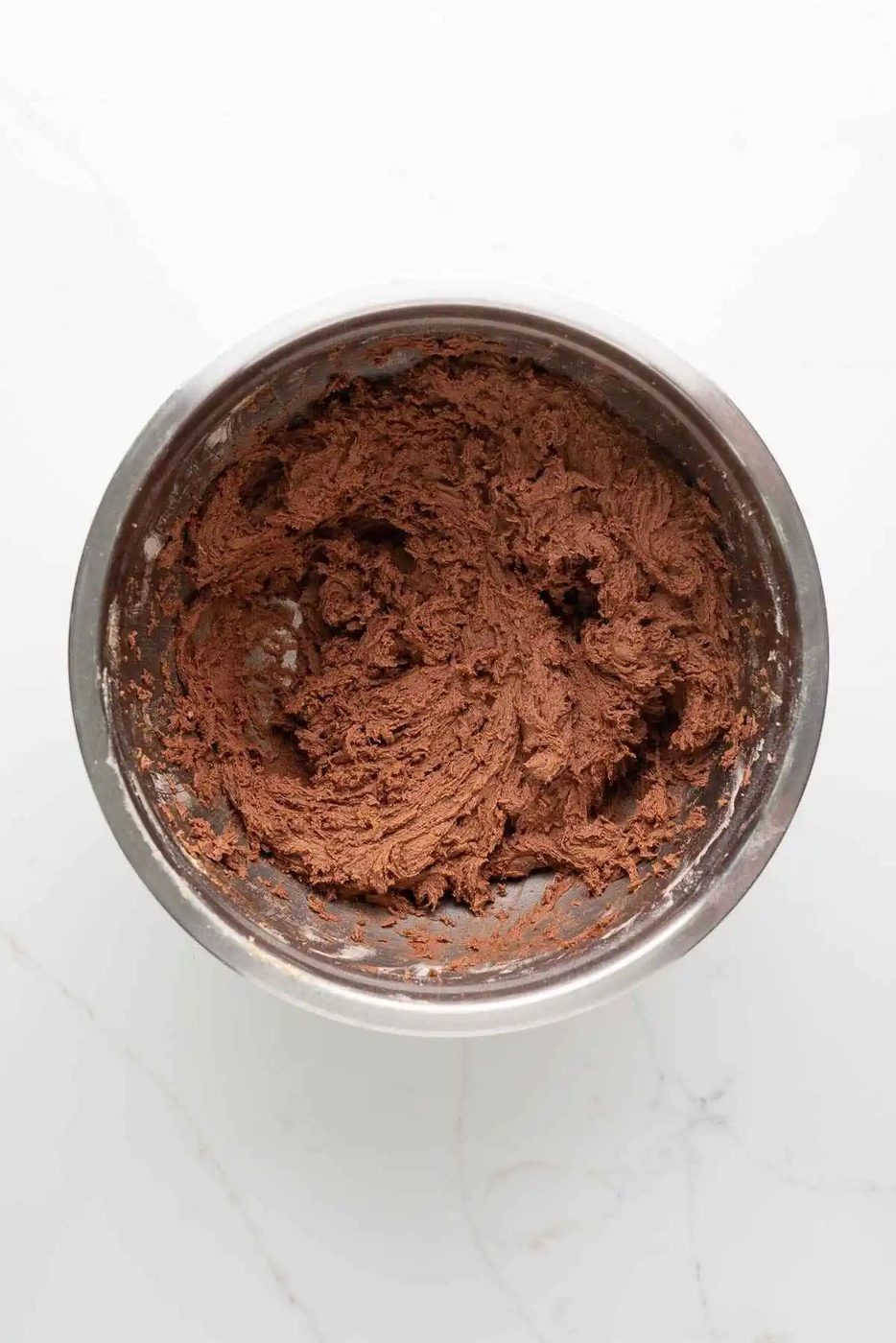 Chocolate cookie dough in a stainless steel mixing bowl on a white marbled background.