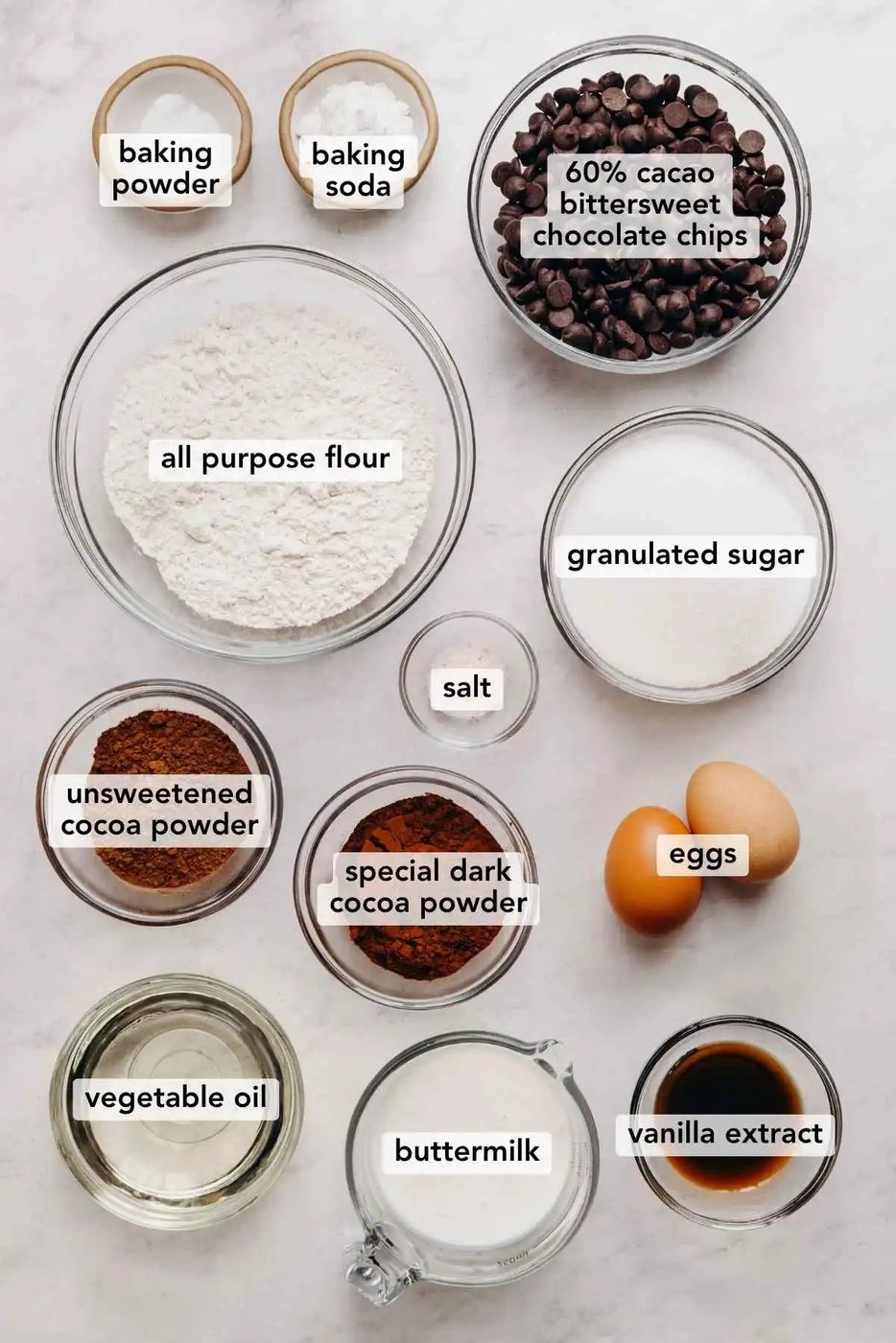 Ingredients in various size bowls labeled as baking powder, baking soda, 60 percent cacao bittersweet chocolate chips, all purpose flour, granulated sugar, salt, unsweetened cocoa powder, special dark cocoa powder, eggs, vegetable oil, buttermilk, and vanilla extract on a white marbled background.