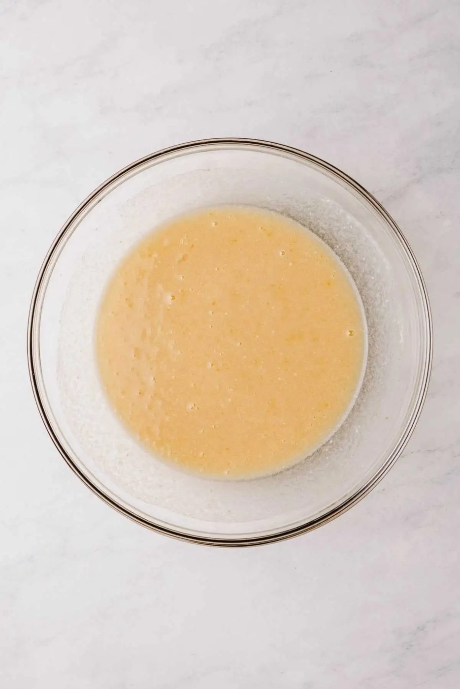 Whisked eggs, sugar, vegetable oil, vanilla extract, and buttermilk in a large glass mixing bowl on a white marbled background.