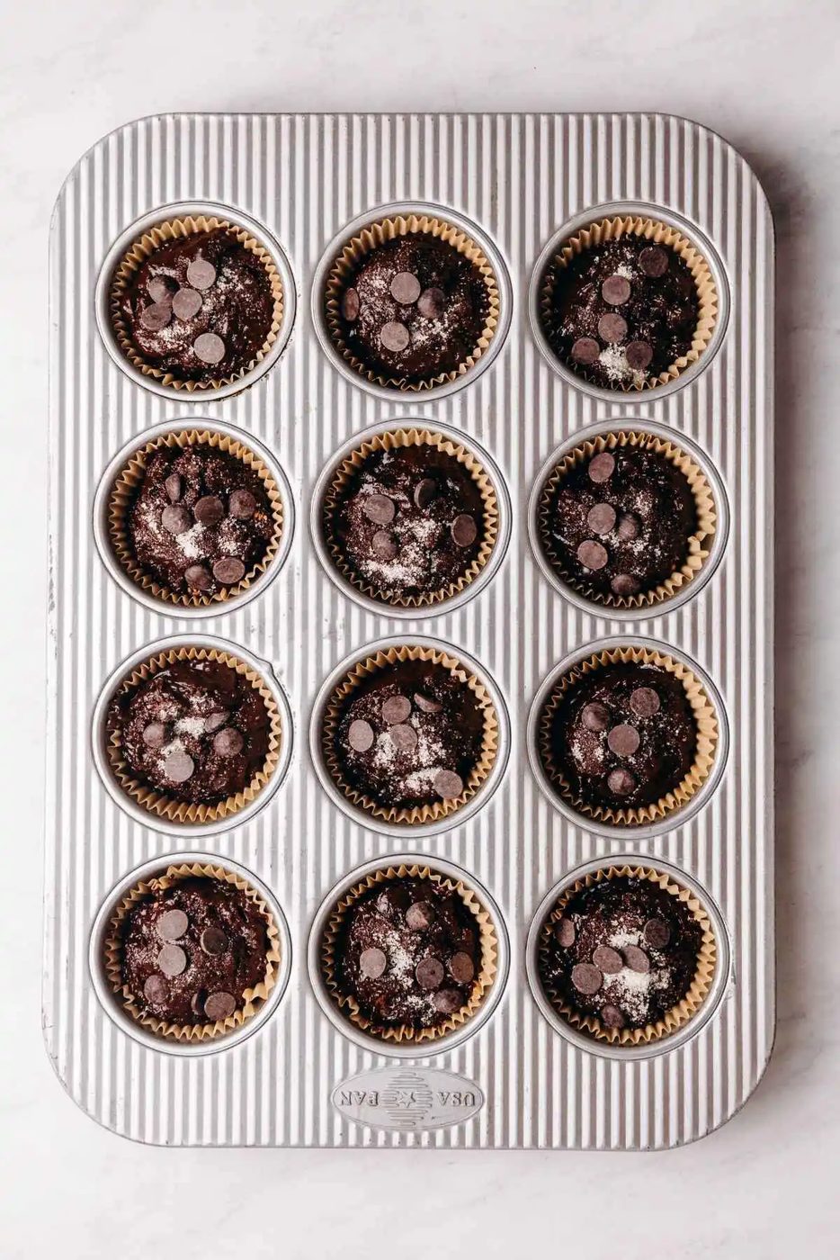 Double chocolate muffin batter topped with sparkling sugar and chunks of chocolate in parchment paper lined metal muffin tin.
