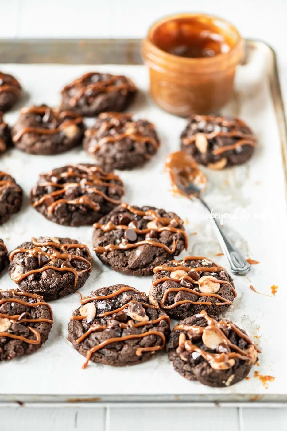 Overhead image of just baked double chocolate salted caramel cookies on a baking sheet with a spoon and jar of a caramel sauce resting close by | All Images © Sovuh