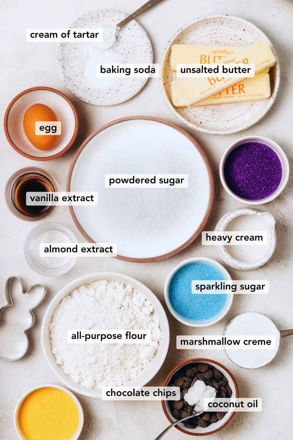 Ingredients in various size bowls and plates labeled as egg, all-purpose flour, unsalted butter, powdered sugar, baking powder, egg, cream of tartar, baking soda, vanilla extract, almond extract, sparkling sugar, heavy cream, marshmallow creme, chocolate chips, and coconut oil on a white marbled background.