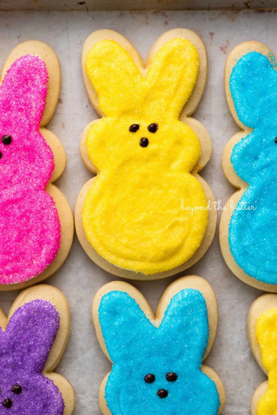 Easter bunny cut out sugar cookies topped with frosting and colors of pink, yellow, blue, and purple sanding sugars on a parchment paper lined baking sheet.