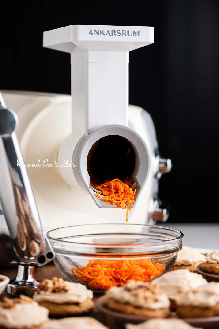 Light creme color ankarsrum mixer using the vegetable cutter attachment to grate carrots into a glass bowl for carrot cake cookies against a dark background.