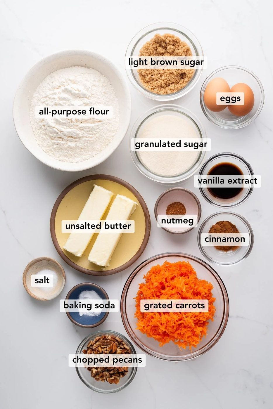 Carrot cake cookies ingredients in various size bowls labeled as all-purpose flour, baking soda, cinnamon, nutmeg, salt, unsalted butter, light brown sugar, granulated sugar, eggs, vanilla extract, grated carrots, and chopped pecans on a white marbled background.
