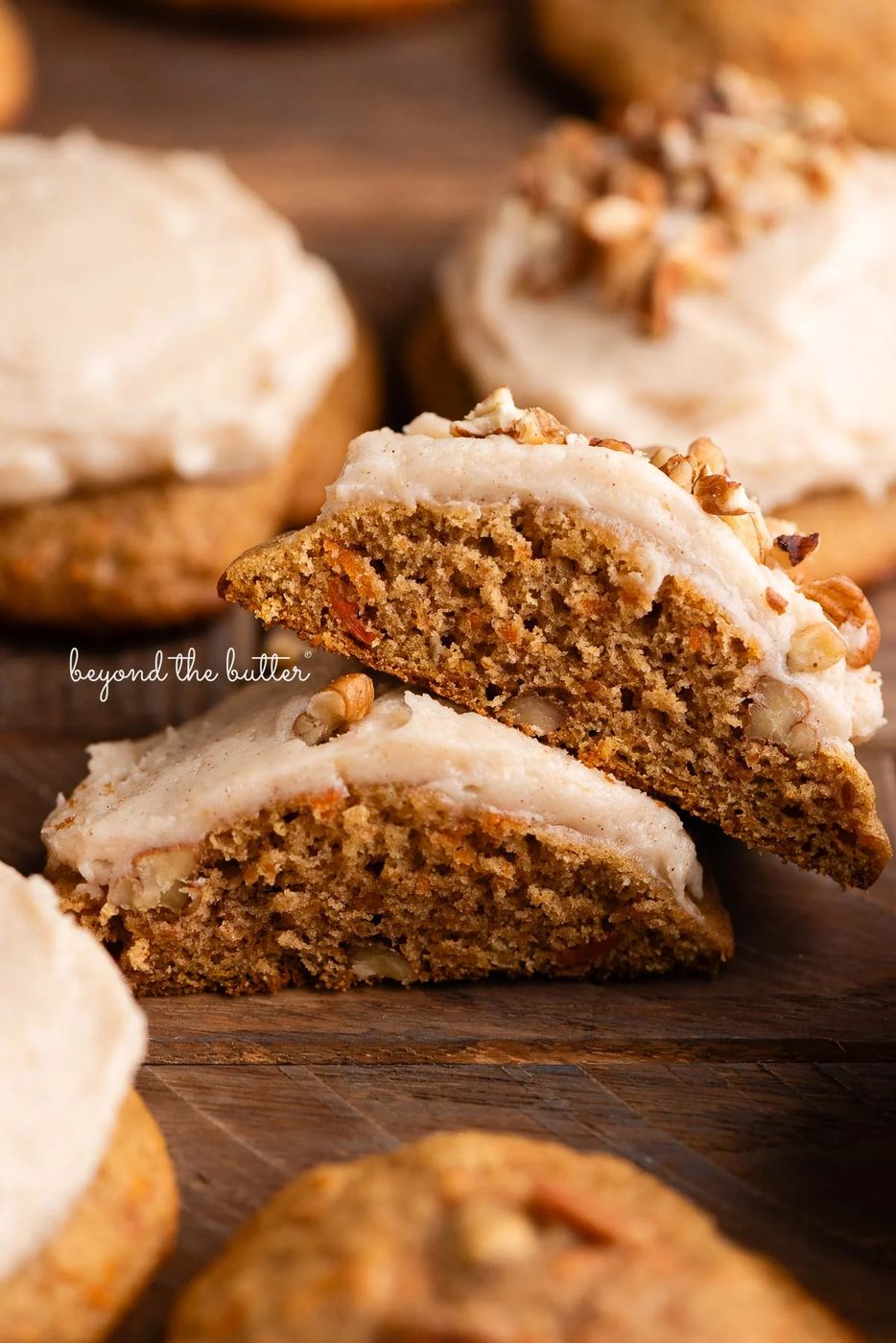 Brown butter cream cheese frosted and unfrosted carrot cake cookies with a sprinkling of chopped pecans around some of the cookies on a dark wood background.