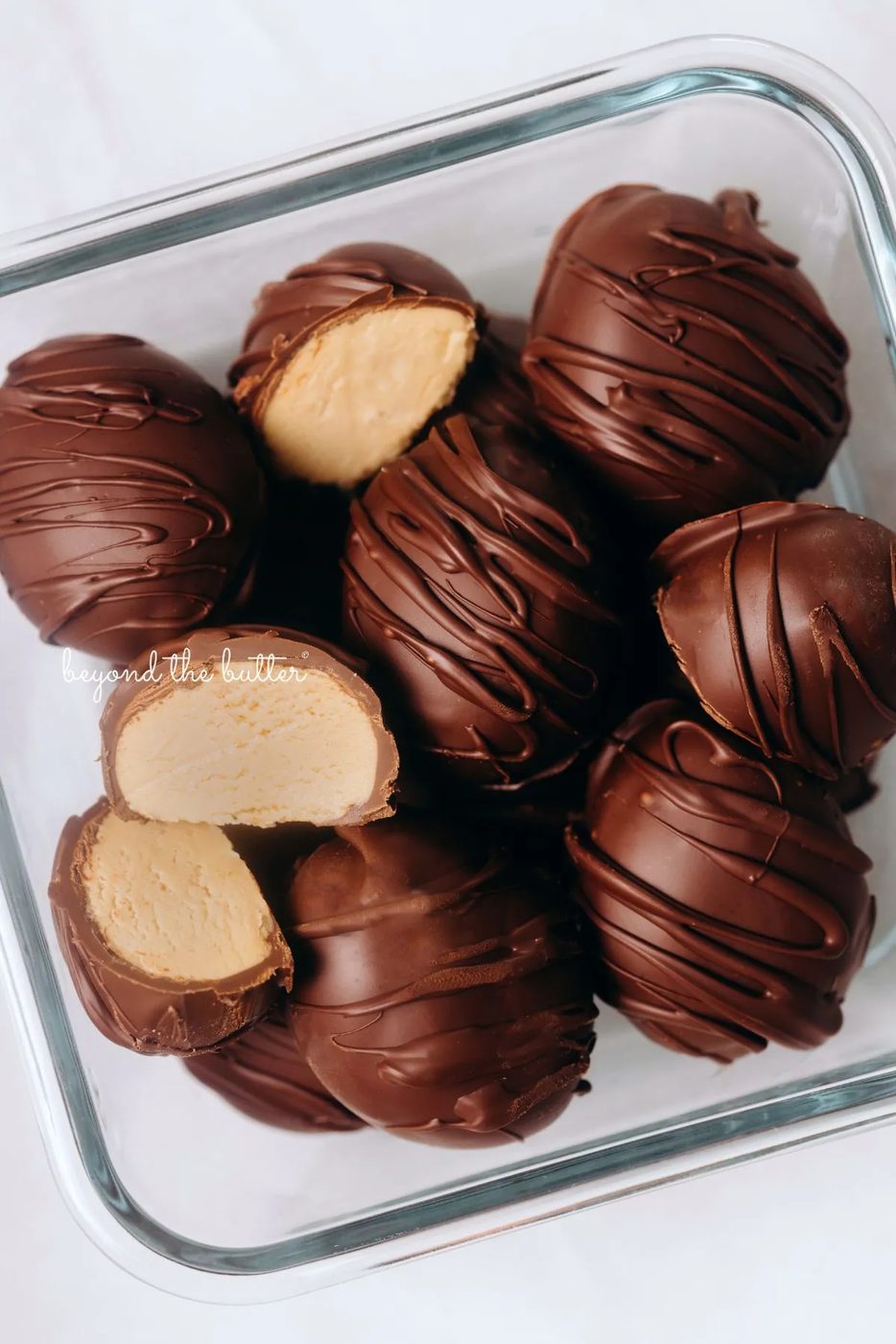 Chilled chocolate covered peanut butter eggs drizzled with more chocolate randomly placed in a glass container with two cut in half on white background.