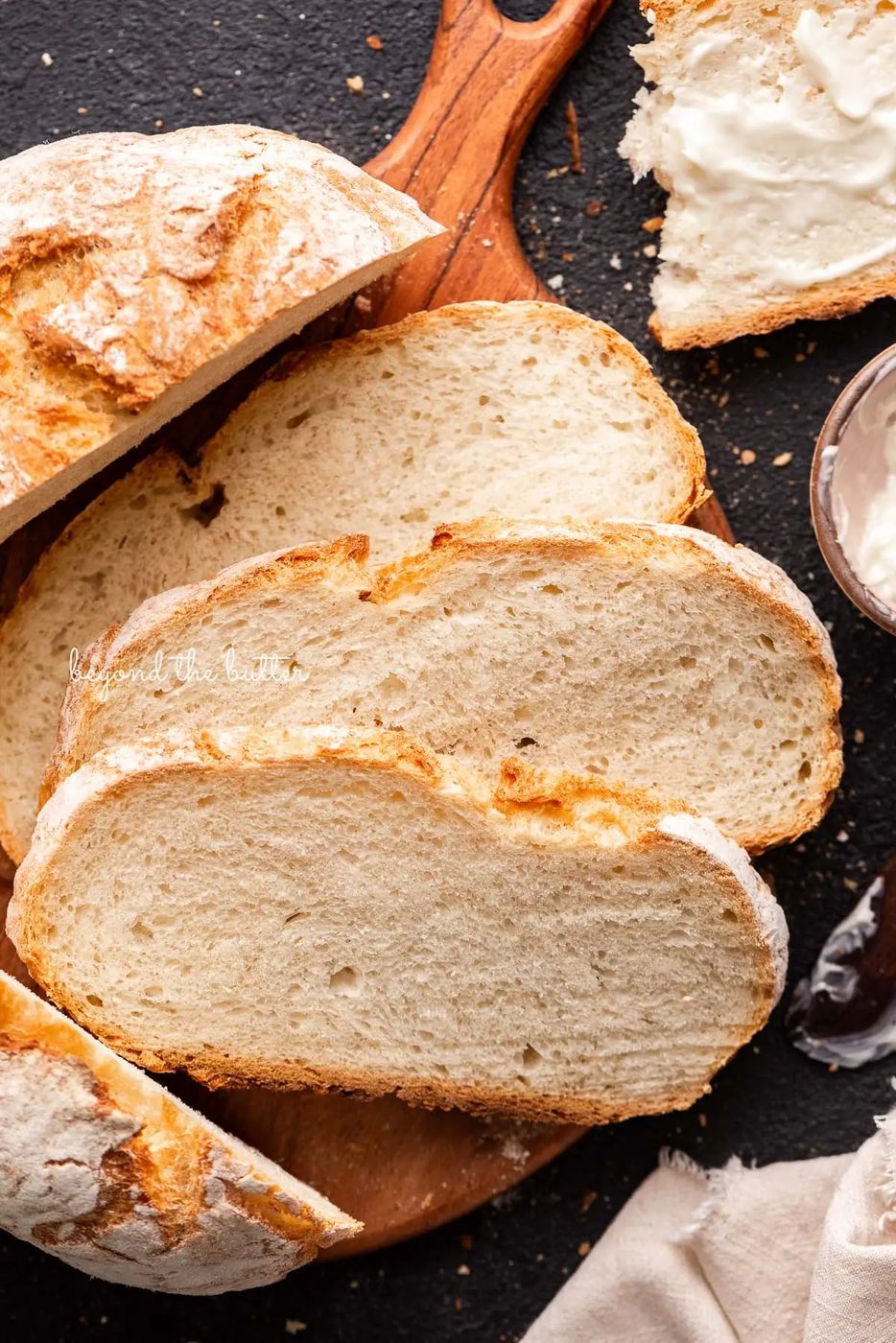 Slices of crusty fresh french bread on a round wooden cutting board next to a small bowl of butter and light beige cloth napkin on a black background.