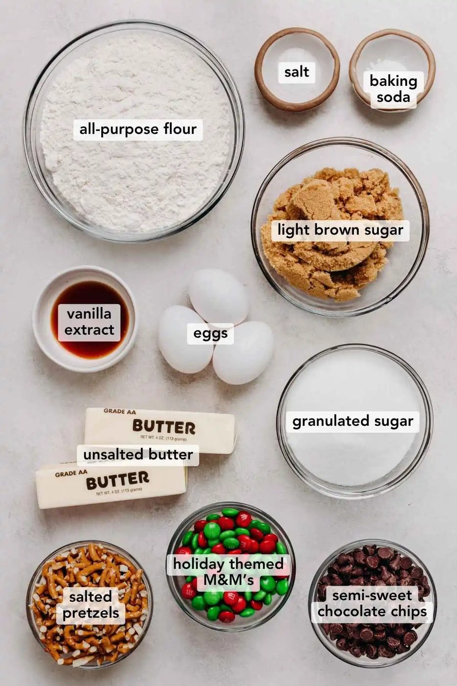 Ingredients in various size bowls labeled as all purpose flour, salt, baking soda, light brown sugar, vanilla extract, eggs, granulated sugar, unsalted butter, salted pretzels, holiday themed M&M candies, and semi sweet chocolate chips on a white marbled background.