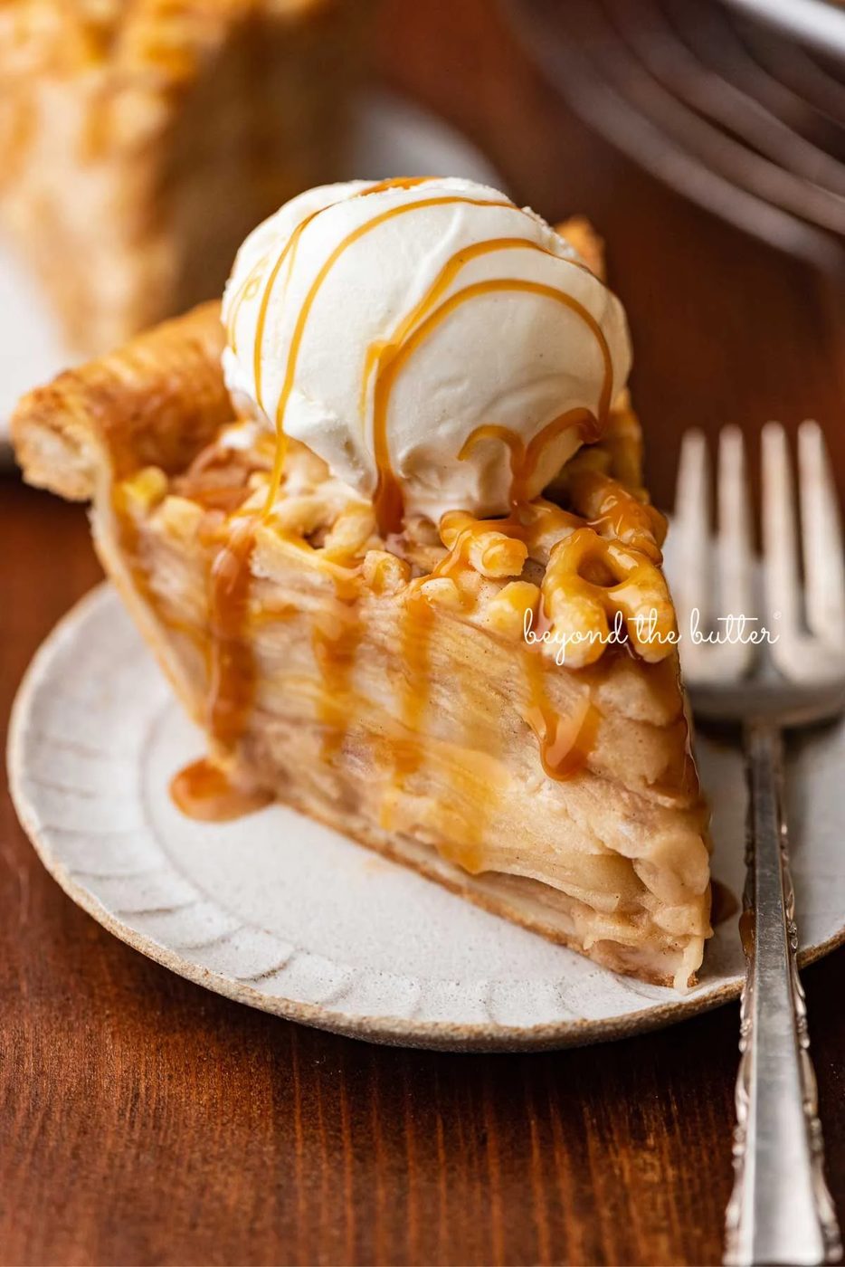 Slice of apple pie drizzled with caramel and topped with a scoop of vanilla bean ice cream on a ceramic dessert plate with a fork placed next it.
