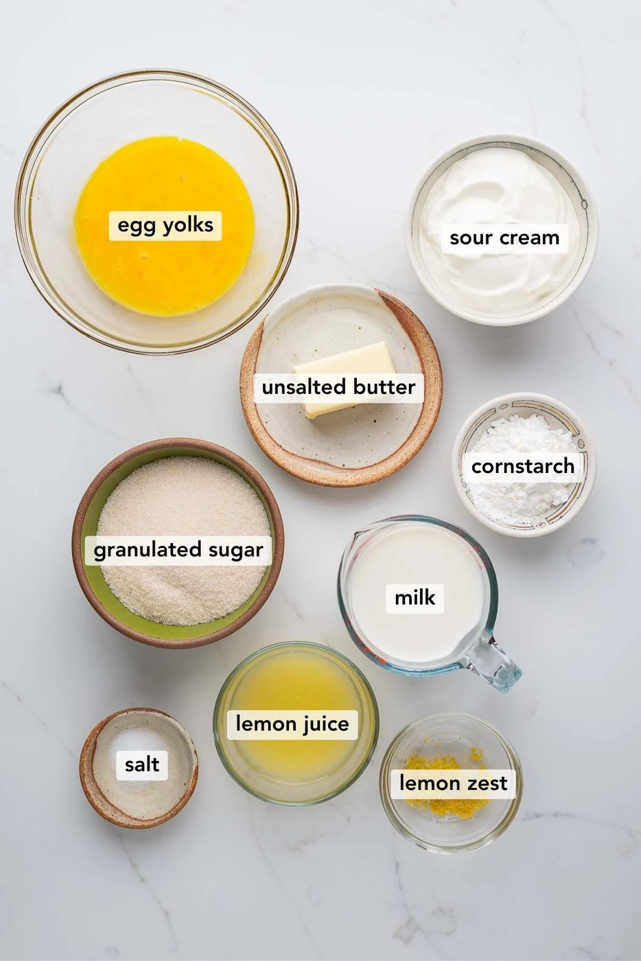 Ingredients in various size bowls labeled as egg yolks, sour cream, unsalted butter, granulated sugar, milk, cornstarch, lemon juice, lemon zest, and salt on a white marbled background.
