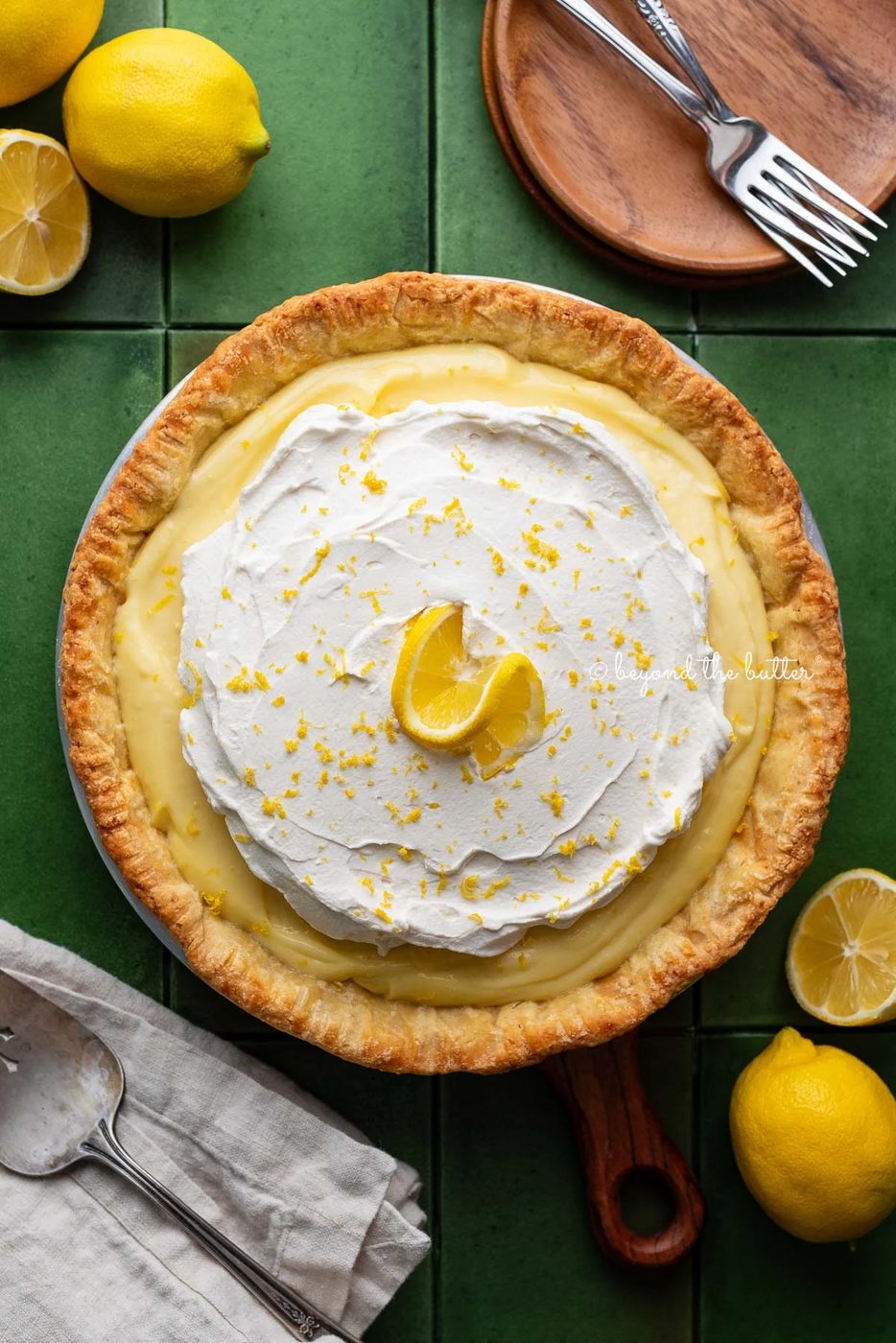 Lemon cream pie topped with fresh whipped cream and lemon zest centered on a green tiled background surrounded by lemons, wood dessert plates, forks, napkin, and pie server.