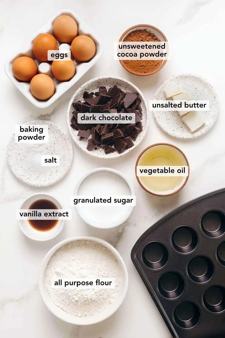 Ingredients in various size bowls labeled as eggs, unsweetened cocoa powder, dark chocolate, salt, baking powder, vegetable oil, vanilla extract, granulated sugar, and all purpose flour on a white marbled background.