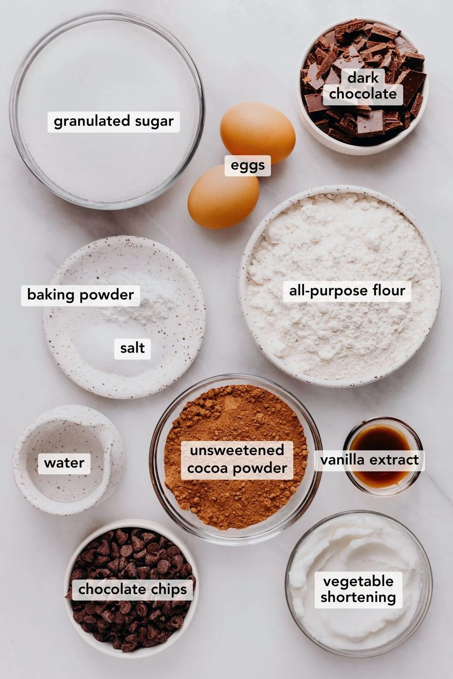 Ingredients in various size bowls labeled as granulated sugar, eggs, dark chocolate, baking powder, salt, all purpose flour, water, unsweetened cocoa powder, vanilla extract, chocolate chips, and vegetable shortening on a white marbled background.