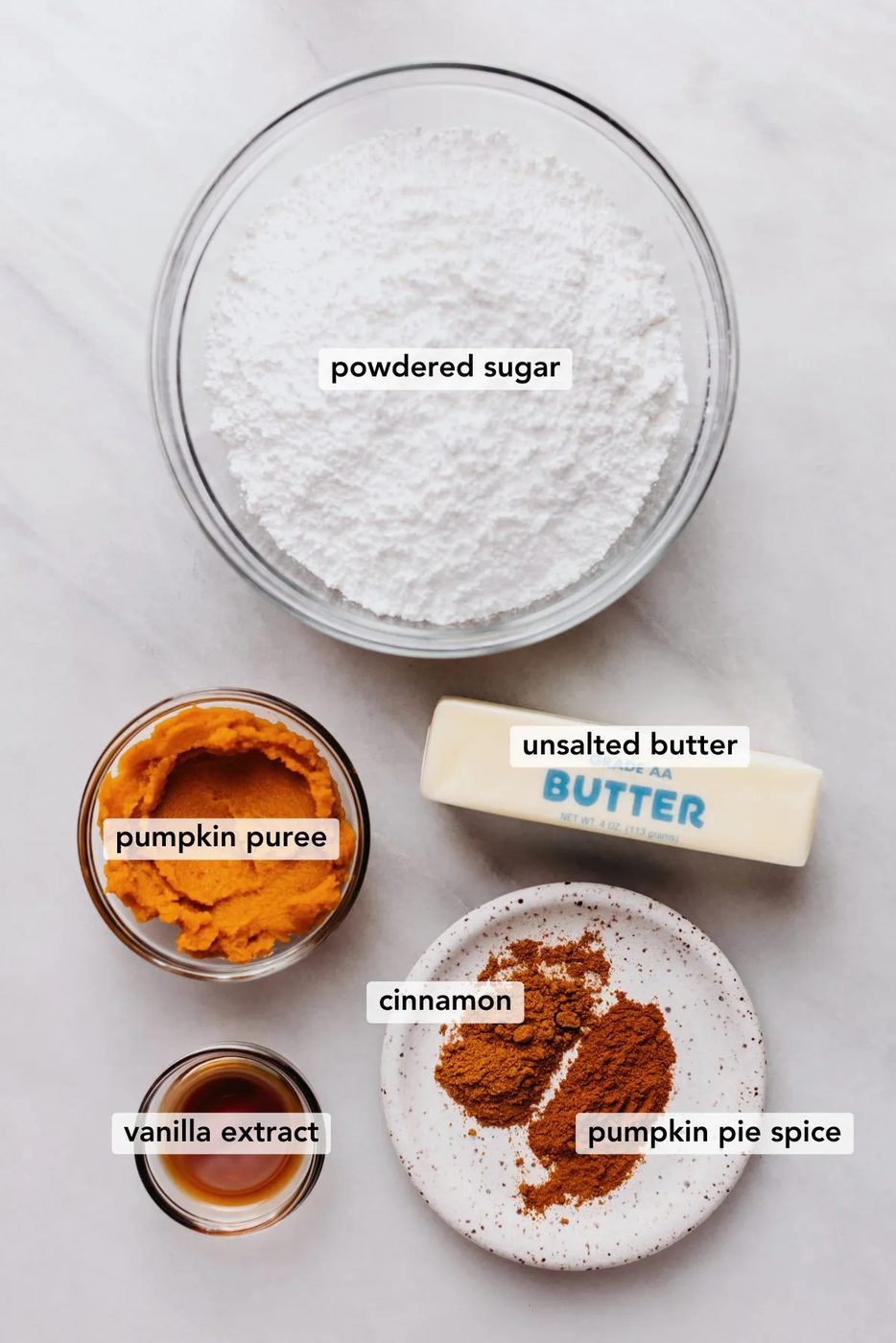 Ingredients in various size bowls labeled as powdered sugar, pumpkin puree, unsalted butter, vanilla extract, cinnamon, and pumpkin pie spice on a white marbled background.