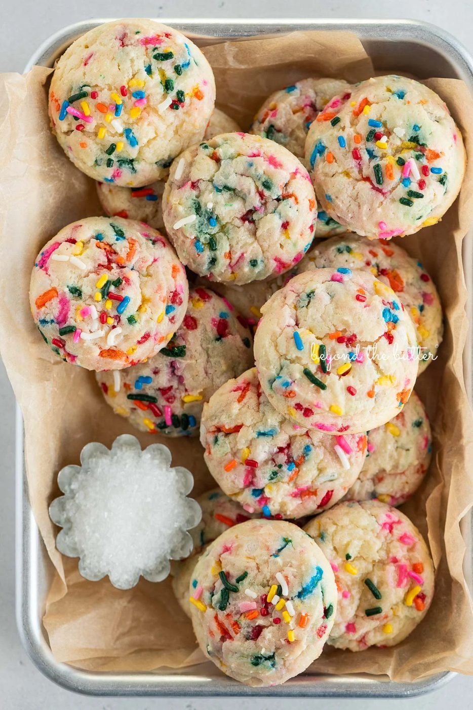 Parchment paper lined small baking sheet filled with funfetti butter cookies and a small scalloped bowl of white sparkling sugar.