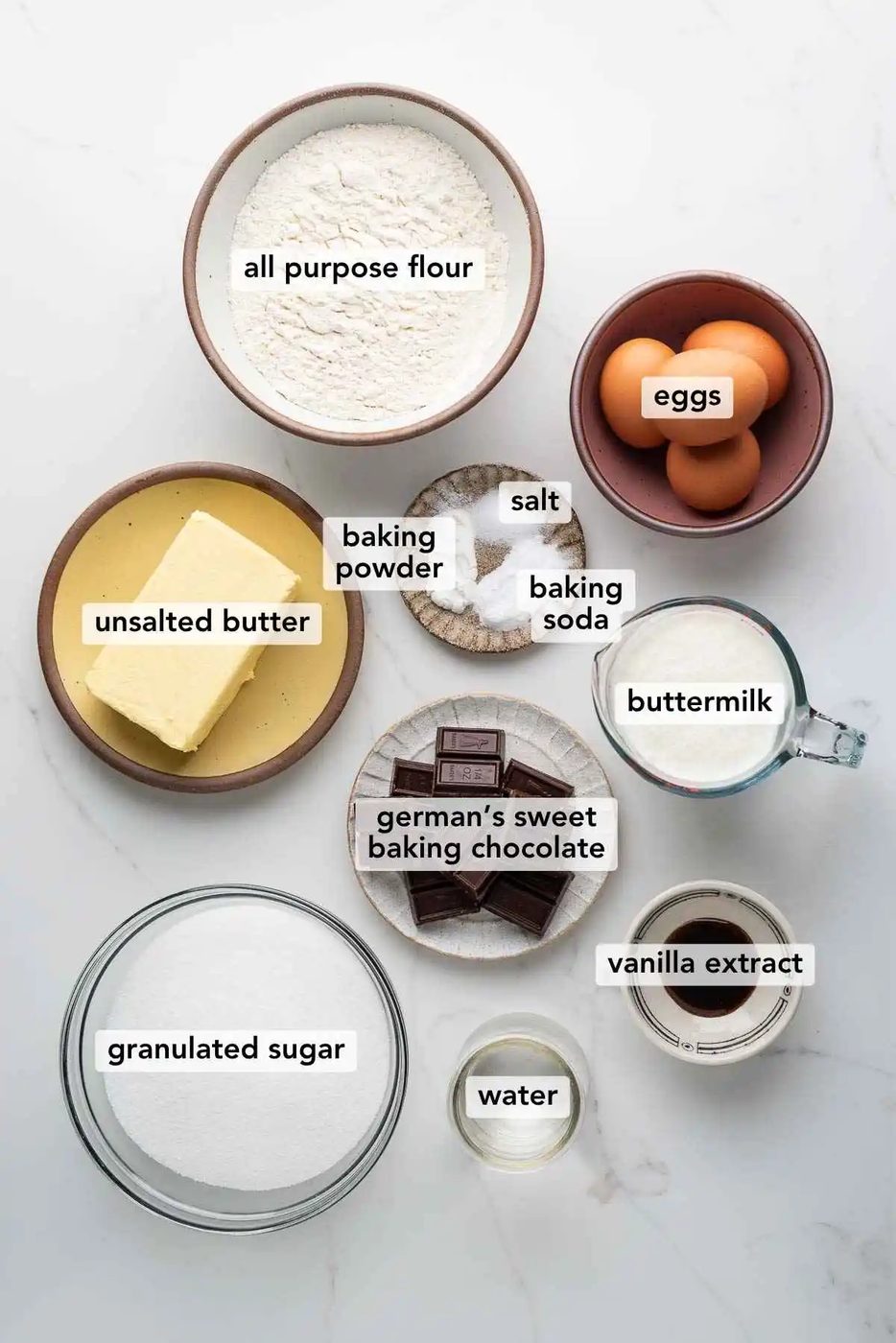 Ingredients in various size bowls labeled as all purpose flour, eggs, unsalted butter, baking powder, baking soda, salt, buttermilk, german's sweet baking chocolate, vanilla extract, water, and granulated sugar on a white marbled background.