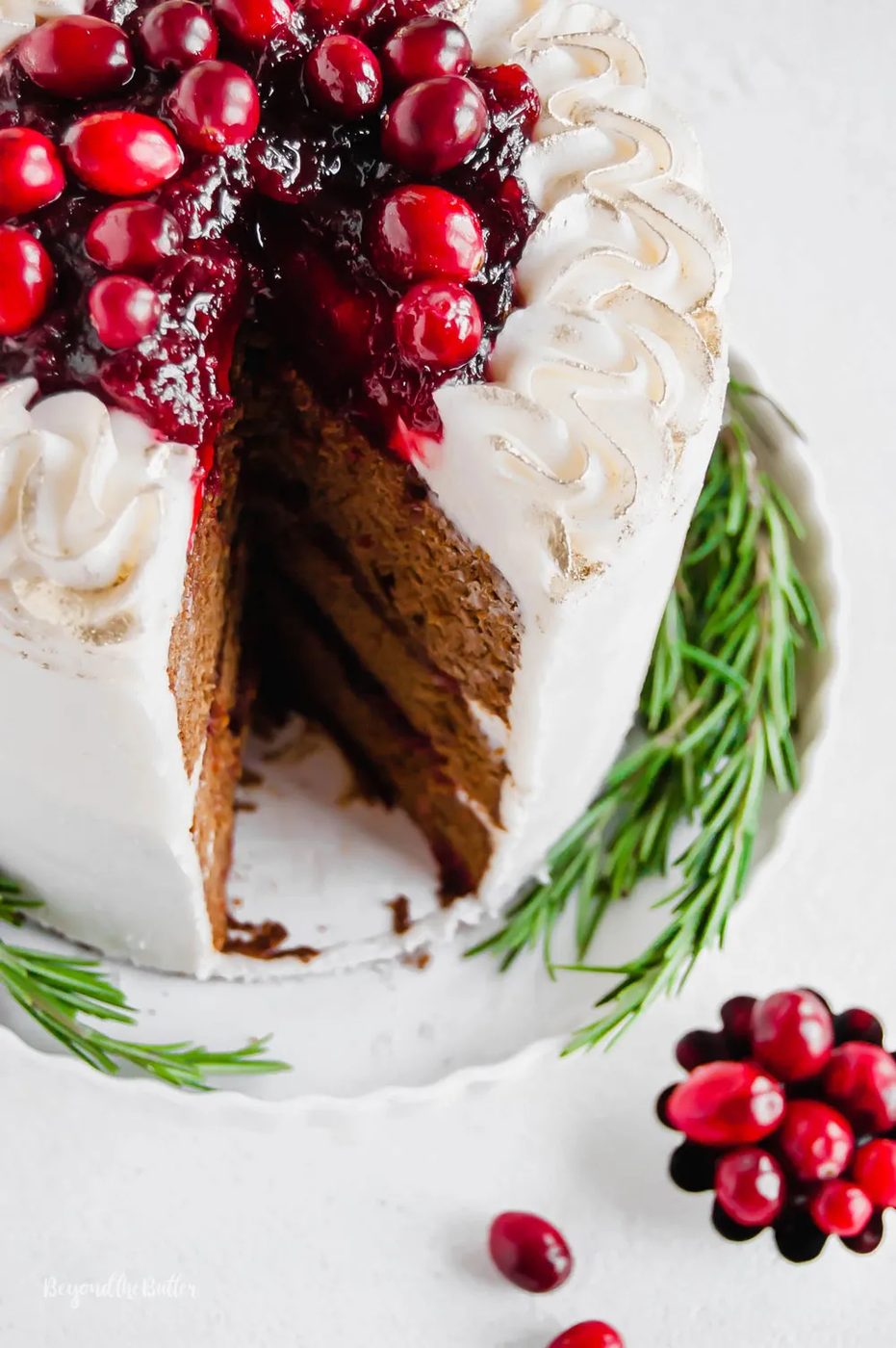 Gingerbread Cake | Angled photo of Gingerbread Cake with Maple Buttercream Frosting and Cranberry Compote with a slice taken out of it and a little bowl of cranberries below it | Image Credit: Sovuh, LLC