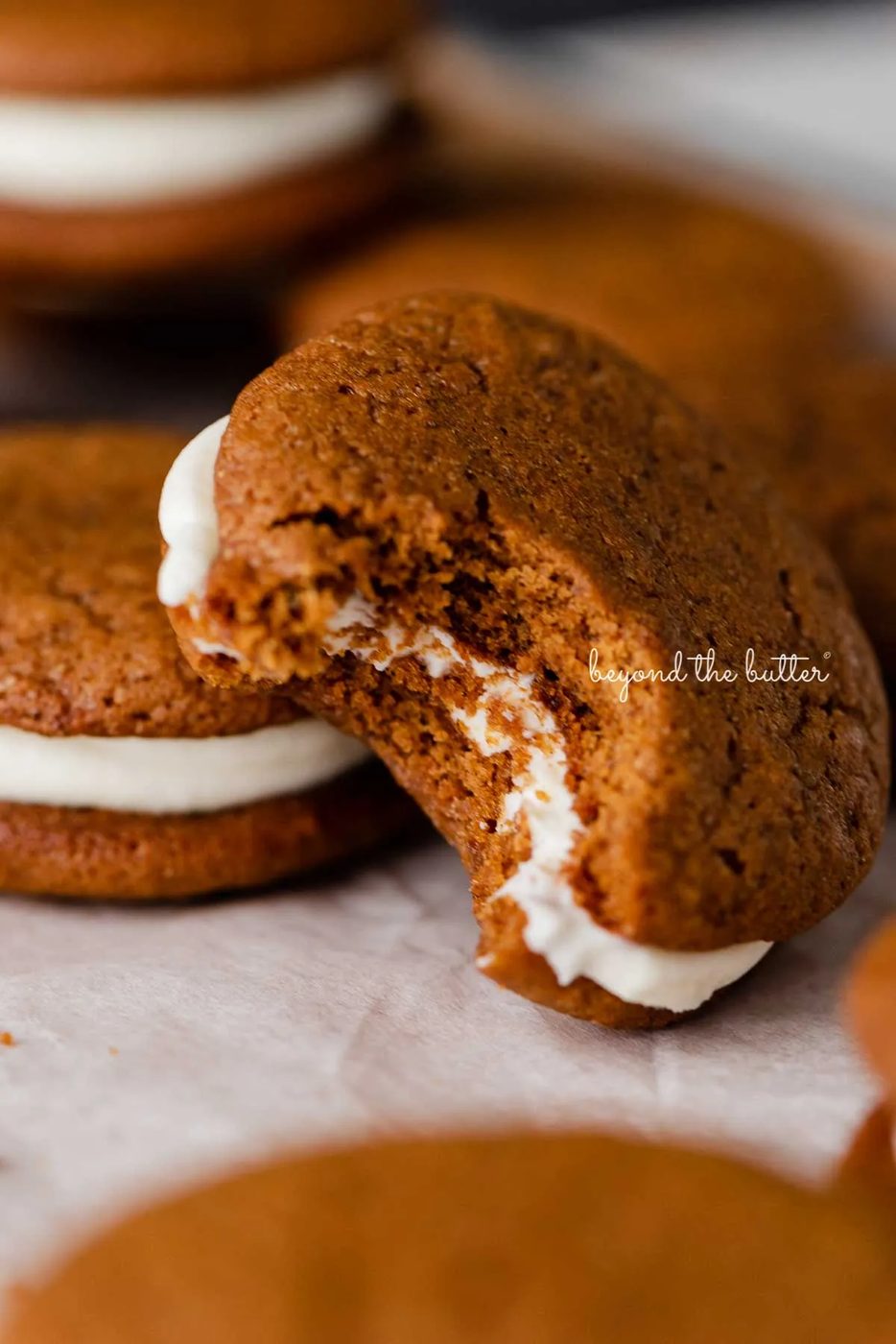 Half eaten house-made gingerbread whoopie pie stacked with more whoopie pies on parchment paper | © Sovuh