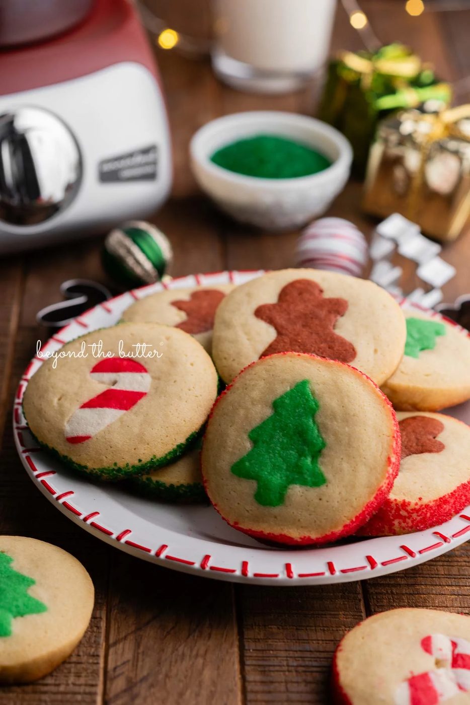A red and white holiday plate full of slice and bake cookies surrounded by more cookies, ornaments, glass of milk, sparkling sugar, and a rustic maroon colored Ankarsrum Assistent Original mixer on a dark wooden background.